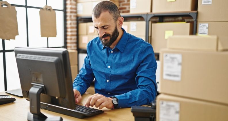 Empreendedor de camisa azul trabalhando no computador em um armazém de loja virtual com caixas.